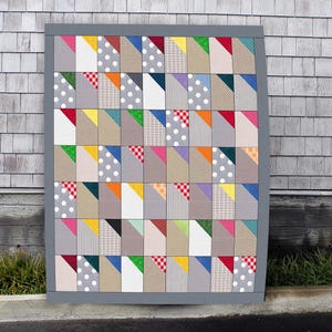 Peut inclure: Une courtepointe patchwork avec une bordure grise. La courtepointe présente un motif répétitif de carrés, chacun divisé en sections de différentes couleurs et motifs, notamment des pois, des rayures et du vichy. La palette de couleurs comprend le rouge, le jaune, le bleu et le gris.