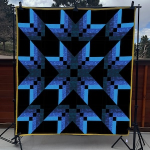 May include: A large, square quilt with a striking geometric star pattern. The design features shades of blue and teal against a black background. The quilt is displayed on a stand, showcasing its intricate patchwork design and craftsmanship.