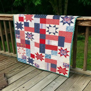 May include: A patriotic quilt with a patchwork design featuring red, white, and blue fabrics. The quilt showcases star patterns and rectangular blocks. It has a green border and is draped over a wooden railing.