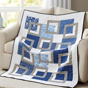 May include: A blue and white quilt with a geometric design. The quilt features squares and rectangles in shades of blue, gray, and white, with a black border. The quilt is draped over a cream-colored sofa.