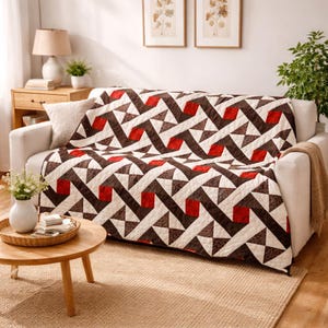 May include: A white sofa is draped with a geometric patterned quilt. The quilt features a repeating design of brown, red, and white shapes. A small wooden side table with a lamp and a vase of flowers are in the foreground. The room has a neutral color palette.