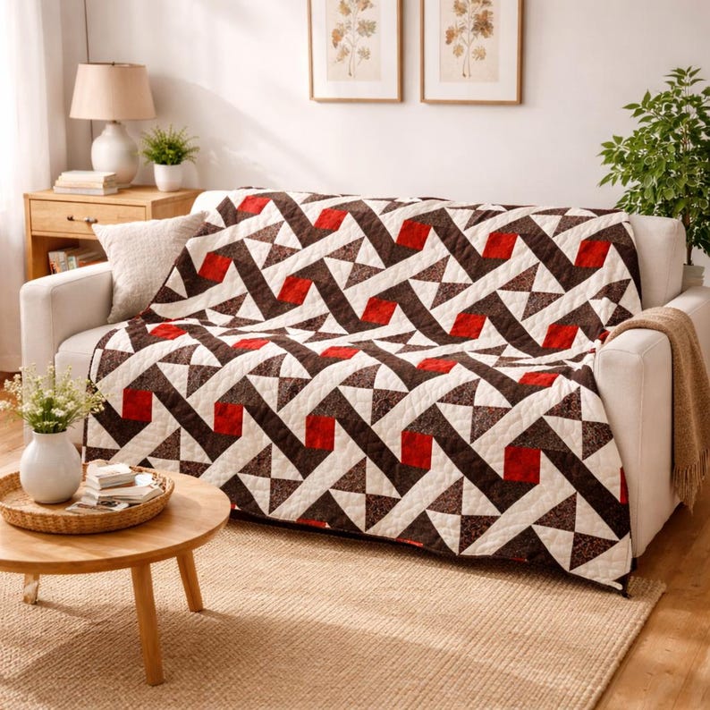 May include: A white sofa is draped with a geometric patterned quilt. The quilt features a repeating design of brown, red, and white shapes. A small wooden side table with a lamp and a vase of flowers are in the foreground. The room has a neutral color palette.