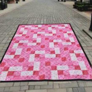 May include: A rectangular quilt featuring a repeating geometric pattern of pink and white fabric squares. The quilt is bordered by a thin black edge and displays various floral and abstract prints within the pink and white sections.