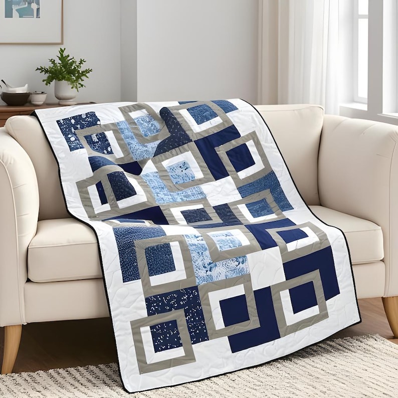 May include: A blue and white quilt with a geometric design draped over a cream-colored sofa. The quilt features squares in various shades of blue, white, and gray, creating a modern, abstract pattern. The quilt's edges are finished with a black border.