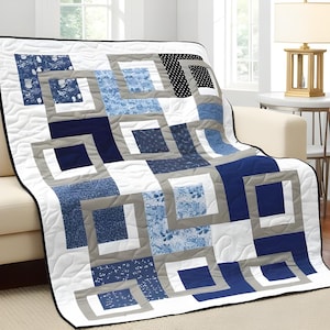 May include: A quilted throw blanket with a geometric design in shades of blue, gray, white, and black. The blanket features a square-in-square pattern, with various blue patterns and solid colors. The blanket is draped over a cream-colored sofa.