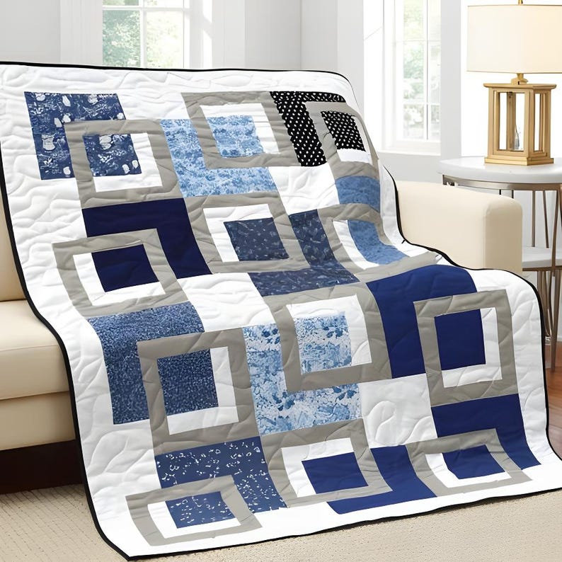 May include: A quilted throw blanket with a geometric design in shades of blue, gray, white, and black. The blanket features a square-in-square pattern, with various blue patterns and solid colors. The blanket is draped over a cream-colored sofa.
