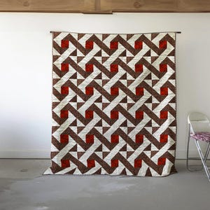 May include: A large quilt with a geometric pattern of brown, white, and red shapes. The quilt is hanging on a wall, showcasing its intricate design. The pattern features zig-zag lines and square and triangular shapes.
