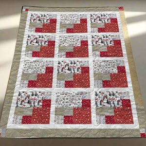 Op de afbeelding: Een rechthoekige quilt met een patchwork-ontwerp. De quilt heeft een raster van vierkanten met afwisselende patronen van rode sterren, winterse taferelen en grijze en witte blokken. De rand is lichtbruin met kleine witte stippen.