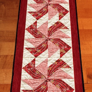 May include: A rectangular quilted table runner with a red and white pinwheel design. The runner features a red border and a black edge. The pinwheel pattern is made of red floral fabric and white fabric.