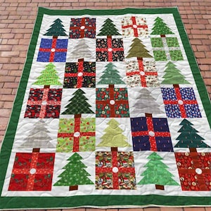 May include: A Christmas-themed quilt with a white background, featuring alternating squares of gift boxes and Christmas trees. The gift boxes are in various colors and patterns, with red ribbon accents. The trees are green, with brown trunks, and the quilt has a green border.
