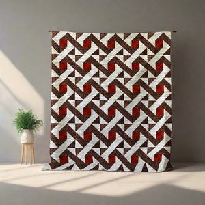 May include: A large quilt with a geometric pattern of white, brown, and red shapes. The quilt is hanging against a neutral wall, showcasing its intricate design. The pattern features interlocking shapes and triangles.