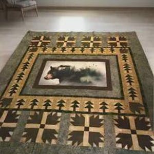 Puede incluir: Una colcha rectangular que presenta un panel central con una ilustración de un oso negro rodeada por un borde de motivos de pinos. La colcha incorpora patrones geométricos en tonos verde oliva, beige y marrón oscuro, con un diseño repetitivo de huellas de oso.