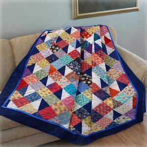 Op de afbeelding: Een patchwork quilt met een verscheidenheid aan kleurrijke bloemrijke en geometrische stoffen vierkanten in een driehoekig patroon, omrand door een effen donkerblauwe stoffen rand met doorgestikte textuur en een felblauwe bies.