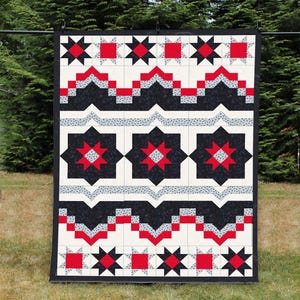 May include: A large, rectangular quilt with a geometric pattern. The quilt features alternating rows of black, red, and white shapes, including stars and squares. The quilt is hanging outdoors, with a green background.