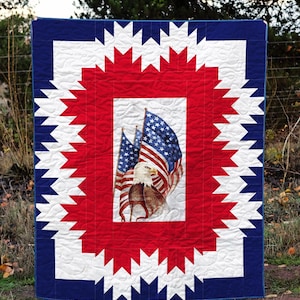 May include: A patriotic quilt featuring a central image of American flags and an eagle. The quilt has a red, white, and blue color scheme with a starburst design around the central image. The border is a solid blue.