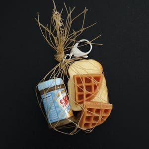 Pode incluir: Um porta-chaves em miniatura com tema de comida, apresentando um pequeno frasco de manteiga de amendoim, duas metades de waffle e uma fatia de torrada, tudo preso numa rede. O porta-chaves tem um fecho prateado e está sobre um fundo preto.