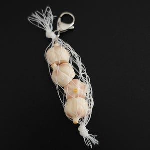 Może przedstawiać: Mała, biała siatka wypełniona kilkoma główkami czosnku. Siatka jest zabezpieczona białą nitką i srebrnym zapięciem. Główki czosnku są w kolorze ecru z odcieniem różu.