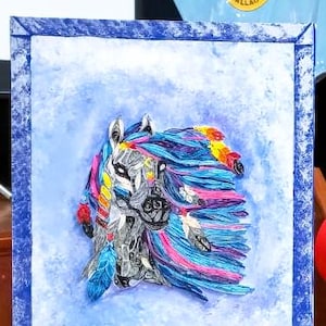 Puede incluir: Obra de arte enmarcada que representa la cabeza de un caballo adornada con plumas coloridas y elementos tribales. La obra utiliza tonos de azul, rosa, amarillo y blanco, con un fondo texturizado y un marco azul.