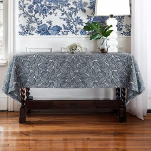 May include: A rectangular tablecloth with a leaf pattern in shades of gray and white covers a dark wooden table. The table is in a room with a floral wallpaper and a white lamp.