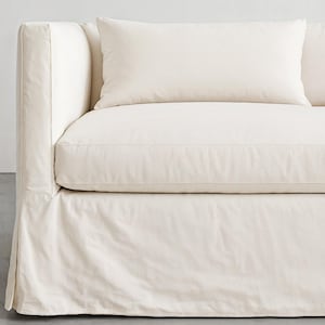 以下が含まれることがあります： 長方形のクッションが付いたクリーム色のソファ。ソファはスカート付きのベースと厚いクッションシートを備えています。アームレストは幅が広く、生地は柔らかくニュートラルな素材のようです。全体的なデザインはシンプルでモダンです。