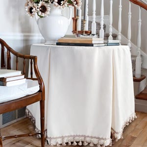May include: A cream-coloured round table with a tassel-trimmed tablecloth. A white vase with sunflowers and a stack of books are on the table. A wooden chair and a staircase are in the background, creating a classic interior scene.