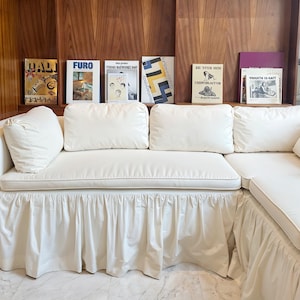 May include: A white sectional sofa with a ruffled skirt, paired with a wooden wall shelf displaying several books. The books have various covers with text and graphics. The sofa has several white cushions.