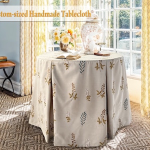 May include: A round, custom-sized handmade tablecloth in a neutral beige colour, adorned with embroidered floral and leaf patterns in shades of gold and blue. The tablecloth is draped over a round table, creating a soft, elegant look. The text "Custom-sized Handmade Tablecloth" is in the image.