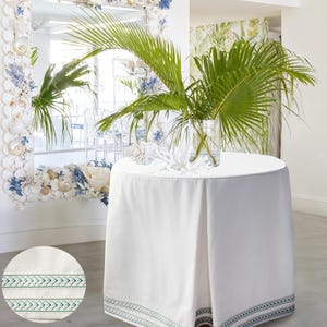 May include: A round white table with a white tablecloth and a green and blue patterned border. A glass vase with palm fronds sits on top. A mirror decorated with shells and flowers is in the background.