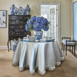Op de afbeelding: Een interieur met een ronde tafel bedekt met een zilveren doek en een franjebeslag. Een grote vaas met blauwe hortensia's staat op de tafel. Een zwarte kast met blauw-witte porseleinen vazen staat op de achtergrond.