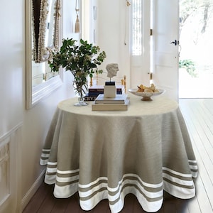 May include: A circular table draped with a beige tablecloth, edged with three white stripes. The table displays books, a small bust, and a fruit bowl. A vase of greenery is also present. The table is approximately 120 cm in diameter.