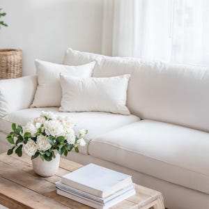 Op de afbeelding: Een lichte, luchtige woonkamer met een witte hoekbank en drie bijpassende kussens. Een houten salontafel toont een vaas met witte bloemen en een stapel boeken. Een gevlochten mand en een potplant zijn zichtbaar op de achtergrond.