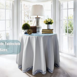 Peut inclure: Une nappe ronde blanche avec une bordure à motifs bleus, drapée sur une table. L'image montre également une lampe, une petite plante, des livres et un vase de fleurs. Le texte à gauche indique "Handmade Tablecloth Custom Sizes."