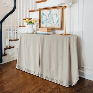 May include: A beige console table with a decorative skirt and a framed artwork. A white vase with yellow tulips, books, and a gold lamp are on the table. The table is set against a white wall and a staircase with a dark blue banister.