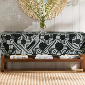 Peut inclure: Une table décorative avec une nappe à motifs noirs et blancs. Une étagère en bois en dessous présente des livres. Un vase de fleurs blanches est posé sur la table et un miroir décoratif est accroché au mur.