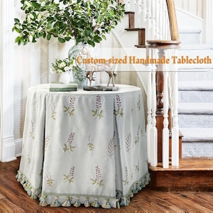 May include: A round, custom-sized handmade tablecloth in a light blue hue, adorned with embroidered floral patterns in purple and green. The tablecloth features a decorative fringe with tassels. The text "Custom-sized Handmade Tablecloth" is visible.