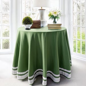 May include: A round, green tablecloth with white stripes at the bottom edge. The table is set with a white lamp, a small topiary, a wooden box, a vase of flowers, and a stack of books. The table is in front of a window.