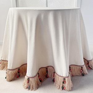 May include: A cream-coloured round tablecloth with a decorative fringe trim. The fringe is beige with red and green accents. The tablecloth is draped over a round table, creating a soft, elegant look. Suitable for special events or home decor.