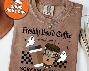 May include: A brown Comfort Colors t-shirt with the text "Freshly Bood Coffee served with SCREAM & SUGAR". The design includes coffee cups, ghosts, and a checkered pattern. A necklace is draped across the shirt.