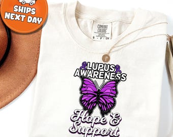 May include: Cream-colored t-shirt with a purple butterfly design and the words "Lupus Awareness" and "Hope & Support". The butterfly has black outlines and a gradient of purple hues. A gold necklace is draped across the shirt.