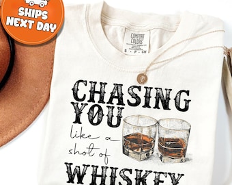 May include: Cream-colored t-shirt with the phrase "CHASING YOU like a shot of WHISKEY" in black lettering. The design includes two whiskey glasses. A gold necklace rests on the shirt.