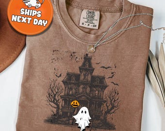 May include: Brown Comfort Colors t-shirt featuring a Halloween design. The graphic includes a haunted house, bats, trees, a ghost, and a jack-o'-lantern. A silver necklace with a pendant is draped across the shirt. The shirt has a crew neck.