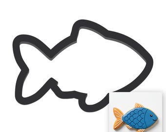 Puede incluir: Un cortador de galletas de metal negro con forma de pez junto a una galleta de pez decorada. La galleta tiene un cuerpo azul, aletas naranjas y una cara sonriente. El cortador es un contorno simple de la forma del pez.