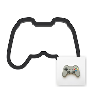 May include: A black outline of a video game controller shape. In the bottom right corner, a grey cookie shaped like a video game controller features a black directional pad, a central button with the letter P, and four coloured action buttons.