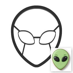 May include: A black plastic cookie cutter shaped like an alien head with large oval eyes. In the bottom right corner, a finished light green cookie with black icing eyes and small curved eyebrows demonstrates the final shape of the cutter.
