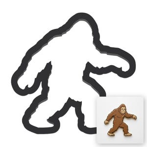 Pode incluir: Um contorno preto de uma figura de Bigfoot com um interior branco. Uma pequena imagem quadrada no canto inferior direito mostra uma figura de Bigfoot marrom com um rosto sorridente e braços estendidos, em um fundo branco.