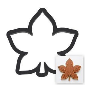 May include: A dark gray leaf-shaped cookie cutter with a thick outline. In the bottom right corner, a small inset shows a brown leaf-shaped cookie with embossed vein details, matching the shape of the cutter, displayed on a white background.