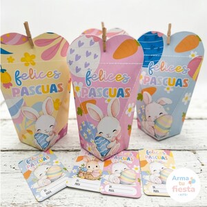 Puede incluir: Cajas de regalo y tarjetas con temática de Pascua. Las cajas tienen forma de corazón en la parte superior, con ilustraciones de conejitos, huevos de Pascua y el texto "felices PASCUAS". Las tarjetas también tienen diseños de conejitos y huevos, con espacio para escribir "DE PARA".