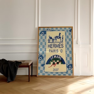 Può includere: Un arazzo tessile rettangolare incorniciato con un motivo a scacchi blu e bianchi e piccoli motivi di auto. Il pannello centrale mostra il testo HERMÈS PARIS, l'icona di una carrozza trainata da cavalli e una grafica LE MANS 2007 in corsivo rosso.