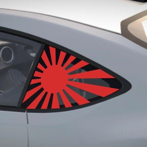 May include: A red rising sun graphic decal on a car window. The design features a central red circle with radiating red lines against a black background. The car is light grey. The decal is a striking visual element.