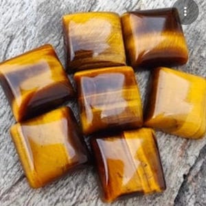 May include: Seven polished tiger's eye stones. The stones are square-shaped with rounded edges. The colours are a mix of golden yellow and brown, with a glossy finish. The stones are arranged on a grey, textured surface.